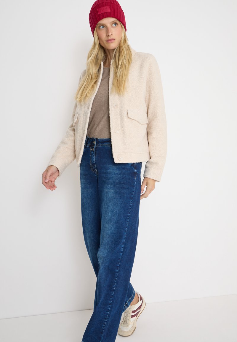 Crème fleece jack met voorzakken, gecombineerd met een lichtbruine top en donkerblauwe wijdleg jeans. Gemaakt met een rode rib gebreide beanie en sneakers.