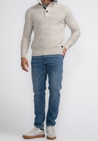 Pull en tricot beige à texture tachetée, col boutonné. Associé à un jean bleu et des baskets de couleur claire. Design simple et décontracté.