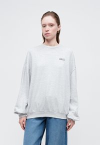 Mujer vistiendo una sudadera oversized de color gris claro con un pequeño logo "Rosie" en el pecho y unos jeans de mezclilla azul holgados, de pie contra un fondo liso.
