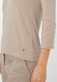 Gestreiftes Langarmoberteil aus beige-weißem Stoff, mit rundem Halsausschnitt und kleinem Logoakzent am Saum. Kombiniert mit beigen Hosen.