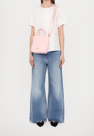 Jasnoróżowa skórzana torebka z podwójnymi uchwytami u góry, regulowanym paskiem i złotym logo. Nosi się ją na białej koszuli i rozkloszowanych niebieskich dżinsach.