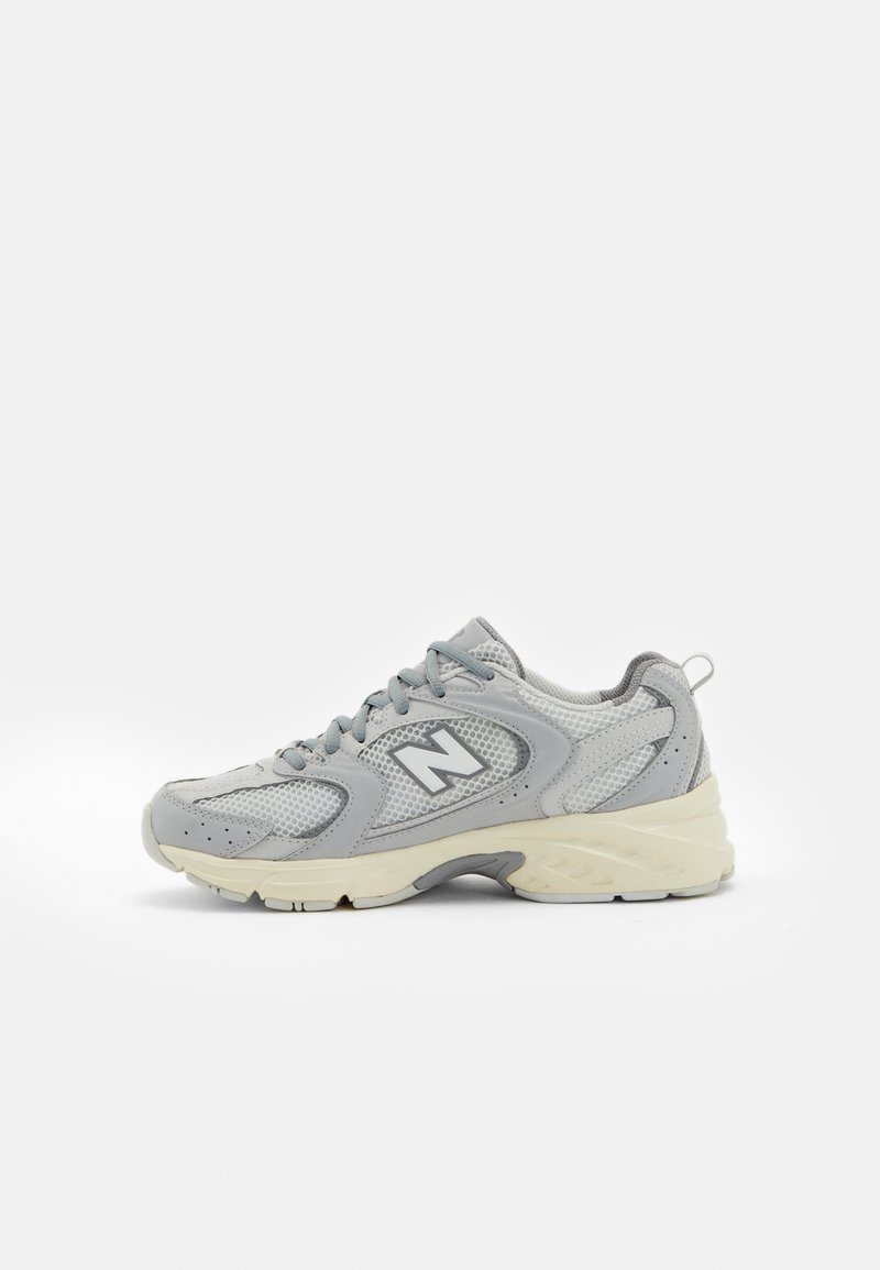 Chaussure de sport gris clair avec une tige en mesh, un col rembourré et un logo, dotée d'une semelle blanche épaisse et de lacets. Design moderne avec des accents texturés.