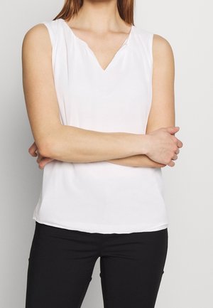 Mujer con los brazos cruzados, vestida con una blusa blanca sin mangas y pantalones negros, sobre un fondo liso y claro.