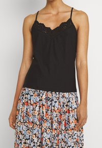 Svart linne med spetsdetaljer och justerbara axelband, i kombination med en plisserad blommig kjol med blå, röda och vita blommor på svart bakgrund.