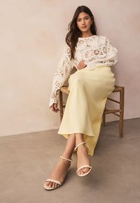 Pull blanc à motif en dentelle avec manches longues, associé à une jupe jaune fluide et des sandales beige à brides à la cheville, assise sur une chaise en bois.