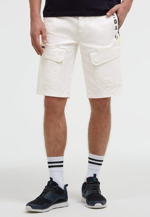 Mann trägt weiße Cargoshorts mit Fronttaschen, weiße Socken mit schwarzen Streifen und schwarze Turnschuhe vor einem einfarbigen Hintergrund.