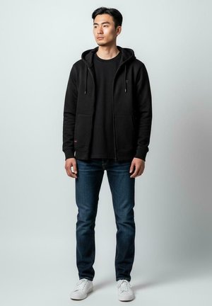 Sweat à capuche noir zippé avec poches frontales, porté sur un t-shirt noir. Associé à un jean bleu et des baskets blanches. Fond uni.