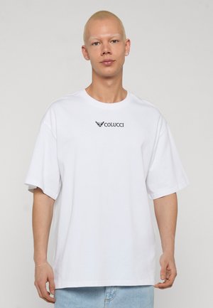 Lühikeste blondide juustega inimene, kes kannab valget üle suurust Colucci T-särki ja helehalli siniseid pükse, seisab ühevärvilise heleda tausta ees.