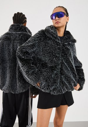 Two women with braided hair wear black and white textured faux fur jackets; one faces forward with purple sunglasses, the other faces away.