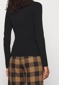 Schwarzes geripptes Langarmshirt kombiniert mit braun-schwarz karierten Hosen, die Textur- und Musterkontraste betonen.