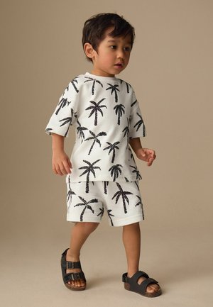 Kleinkind läuft und trägt ein passendes weißes Shirt und Shorts mit schwarzen Palmenmotiven sowie schwarze Sandalen mit Schnalle vor beigem Hintergrund.