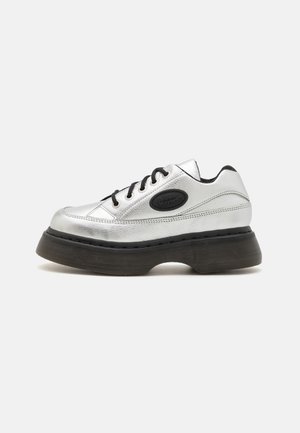 Srebrny skórzany sneaker na platformie z czarnymi sznurowadłami, grubą czarną podeszwą oraz owalną czarną naszywką z logo po boku.