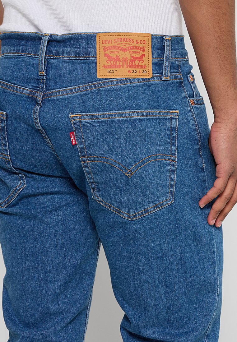 Persona che indossa jeans Levi's 511 blu, con tasca posteriore visibile, etichetta in pelle e mano destra poggiata di lato.