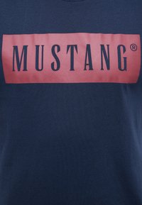 Tamsiai mėlynas medvilnės marškinėliai su dideliu rožiniu stačiakampiu, kuriame rašoma "MUSTANG" ryškiomis juodomis raidėmis. Lygus tekstūra, trumpomis rankovėmis.