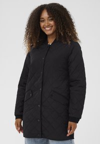 Hymyilevä nainen kiharat hiukset, yllään musta tikattu pitkä takki, jossa nepparit ja kaksi etutaskua, yhdistetty vaaleansinisiin farkkuihin.