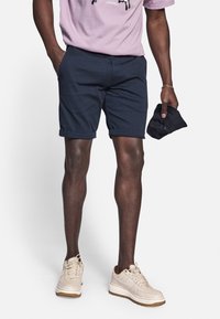 Shorts en coton bleu marine avec un ourlet retroussé, portés avec un t-shirt lilas et des baskets beiges. Une main tient une casquette sombre. Détails de couture visibles.