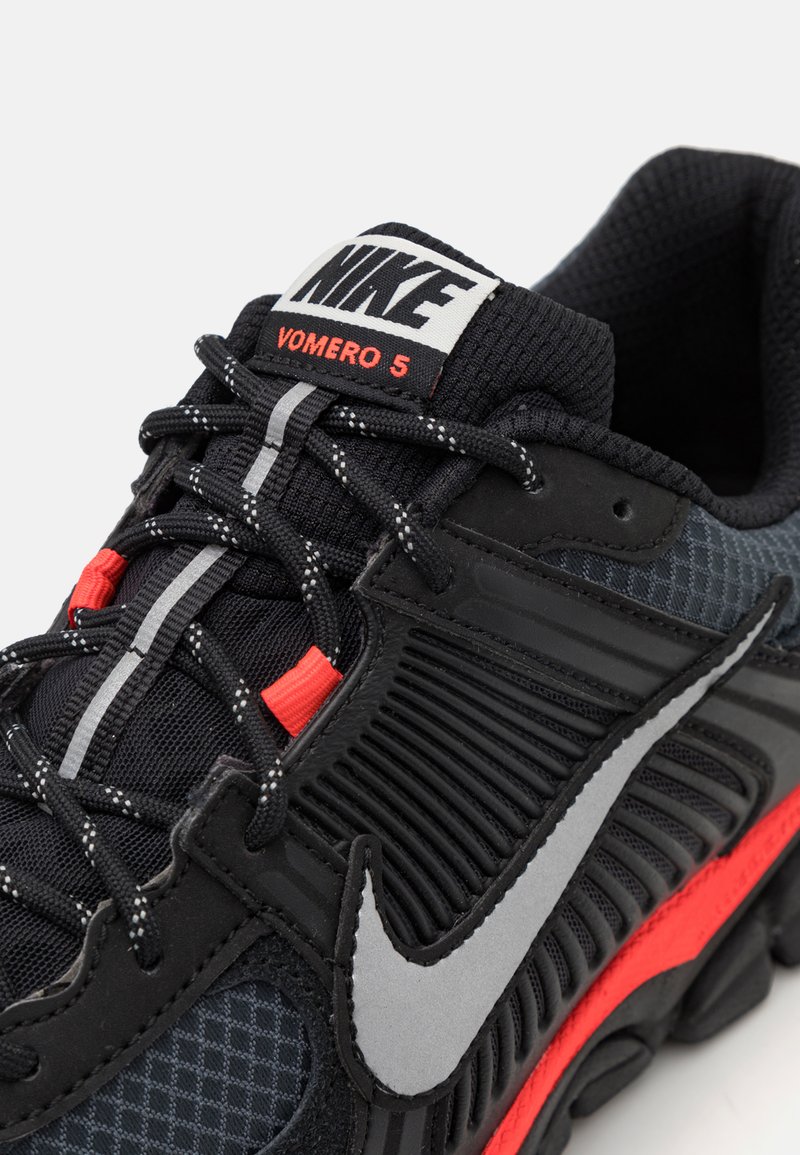 La chaussure de course Nike Vomero 5 présente une tige en mesh noir avec des accents gris, des détails orange et un col rembourré. Semelle texturée avec des rainures de flexion.