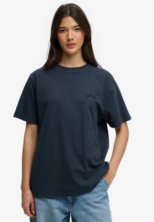 Jonge vrouw met lang donker haar, gekleed in een oversized donkerblauw T-shirt en lichtblauwe spijkerbroek, staand met één hand in haar zak.