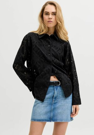 Femme aux longs cheveux blonds porte une chemise noire en dentelle boutonnée et une jupe en denim bleue avec une ceinture noire sur un fond clair uni.