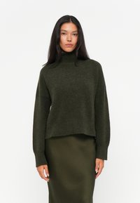 Mørkegrønn strikkegenser med høyt hals, løs passform og lange ermer, kombinert med et matchende satengskjørt. Myk tekstur og minimalistisk design.