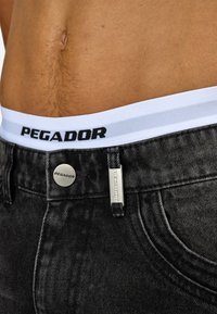 Mustat farkut, joissa on teksturoitu pinta. Valkoinen vyötärönauha, jossa on mustalla kirjoitettu "PEGADOR". Metallinappi ja hopea merkkiyksityiskohta.