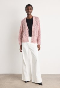 Cardigan rosa lavorato a maglia con una vestibilità ampia, maniche lunghe e tre bottoni. Abbinato a pantaloni bianchi a vita alta e ampia gamba e décolleté neri a punta.
