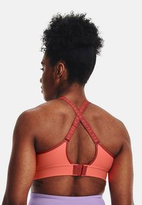 Sporta krūšturis korallkrāsā, izgatavots no gludas auduma. Iezīmēts ar krustu aizmugurē esošām siksnām ar zīmola detaļām, regulējamu aizdari un bezšuvju dizainu.