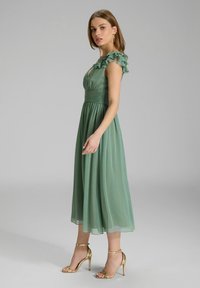Grünes Chiffon-Midi-Kleid mit Rüschenärmeln, tailliertem Oberteil und fließendem Rock. Kombiniert mit goldenen Riemchen-Highheels. Glatte Textur.