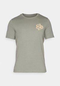 Camiseta de manga corta en verde oliva claro, fabricada en un tejido suave, con un gráfico en color crema que dice "JUST DO IT" en la parte superior izquierda del pecho.