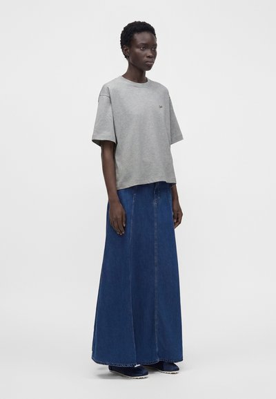 Femme portant un chemisier gris à manches courtes et une longue jupe en jean bleue, debout devant un fond clair uni.