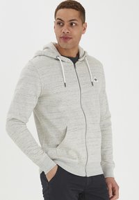 Svijetlosivi zip-up dukserica izrađena od teksturiranog materijala, s prednjim džepom, vezicama i malim logotipom na prsima.