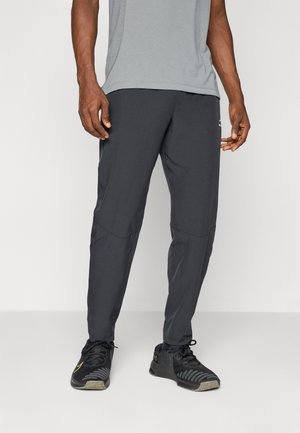 Mann trägt schwarze Sporthose und schwarze Sportschuhe, steht vor einem einfarbigen hellen Hintergrund.