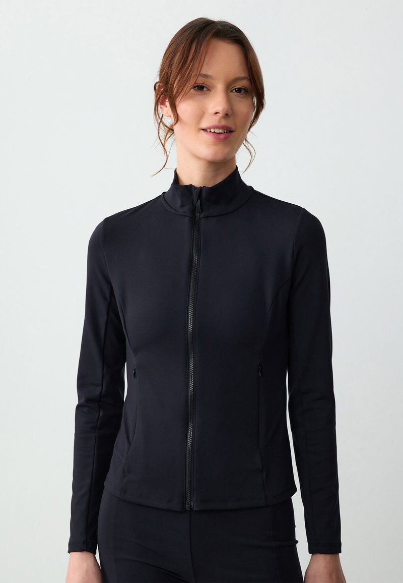 Giacca nera con zip realizzata in un materiale liscio e stretch. Presenta un collo alto, maniche lunghe e tasche laterali. Design aderente.