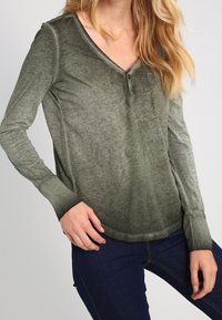 Olivgrünes Langarmshirt mit V-Ausschnitt, ausgestattet mit einer Knopfleiste, einer Brusttasche und einem glatten, leicht strukturierten Stoff.