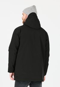 Musta huppari, jossa on sileä pinta, olkapäätaskut ja tasainen helma. Harmaa neulottu pipo tuo kontrastiväriä.