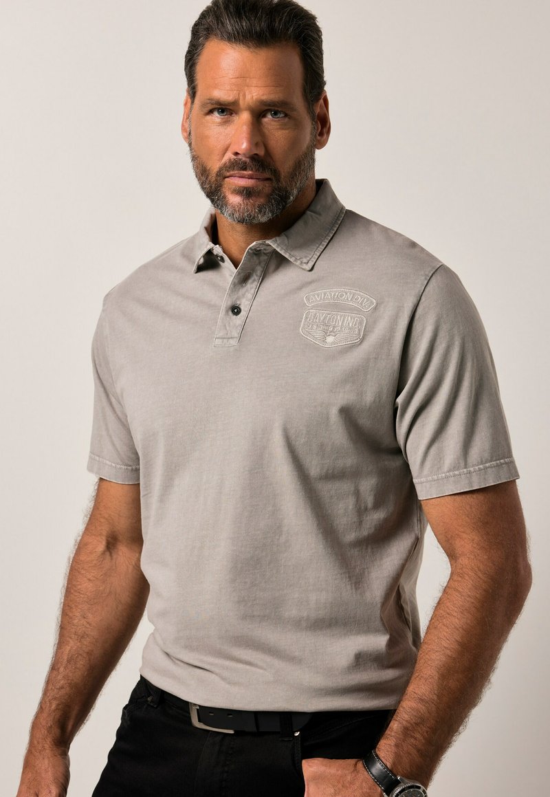 Šedé polo tričko s krátkými rukávy, jemnou texturou a límečkem, s vyraženým logem na levé straně hrudi. Viditelný černý opasek a náramkové hodinky.