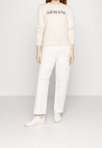 Sudadera rosa claro con logo texturizado, combinada con pantalones de pierna ancha blancos y zapatillas blancas. Tejido suave y corte relajado en toda la prenda.