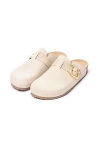 Sabots en suède beige à enfiler avec des orteils ronds, des boucles en or réglables sur les côtés et des semelles texturées marron clair.