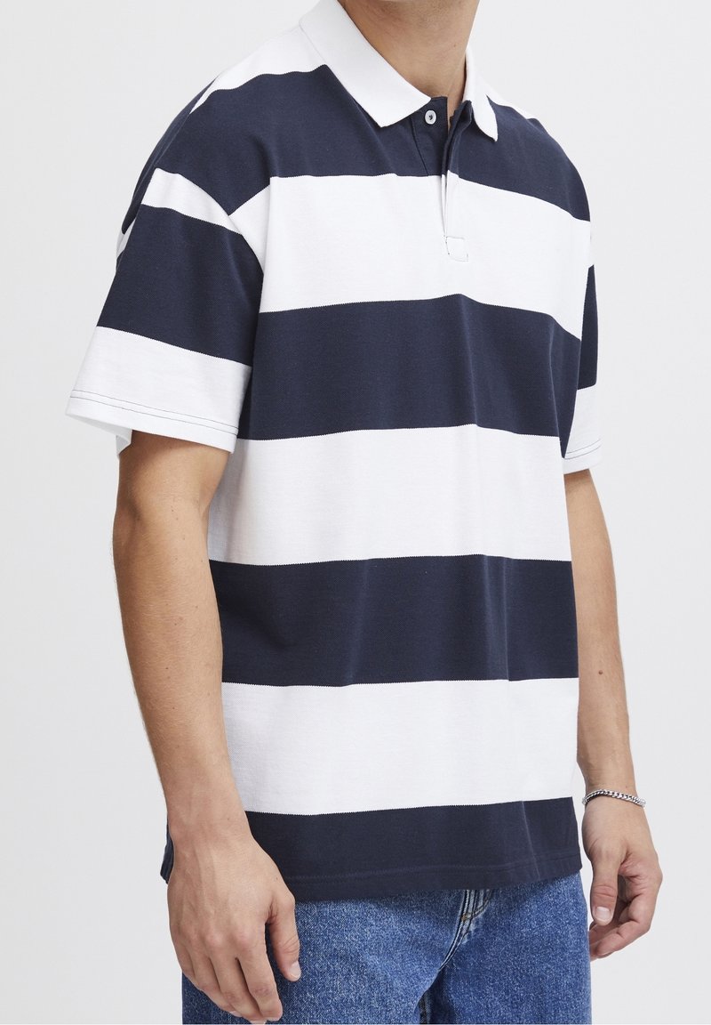 Homme portant un maillot de rugby à manches courtes avec de larges rayures horizontales bleu marine et blanches, un col blanc, et un jean bleu en denim.