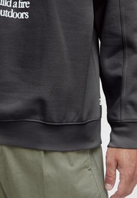 Sudadera gris con puños y dobladillo acanalados, que presenta texto blanco. Mostrada con pantalones verde olivo, destacando la textura y el ajuste de la tela.