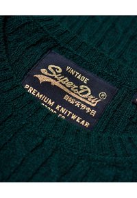 Smaragdgrüner gestrickter Pullover mit gerippter Textur, ausgestattet mit einem dunkelblauen Etikett. Das Etikett zeigt "Superdry" in Gold, was auf hochwertige Strickwaren hinweist.