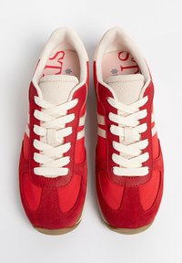 Les baskets rouges présentent un mélange de textures lisses et en daim, des lacets blancs, et trois bandes horizontales de chaque côté. Conception à bout arrondi.