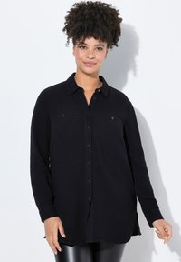 Schwarze Button-Up-Hem mit langen Ärmeln, strukturiertem Stoff, zwei Brusttaschen und einem Kragen. Besitzt eine lockere Passform und seitliche Schlitze.