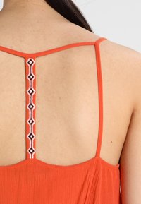 Haut en tissu orange avec des bretelles fines, présentant un détail brodé décoratif au dos. Tissu doux avec des plis subtils.