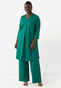 Teal, lång tunika med V-ringning och sidoöppningar, kombinerad med vida byxor. Slät, lättviktsmaterial med en avslappnad passform. Gula sandaletter med remmar.