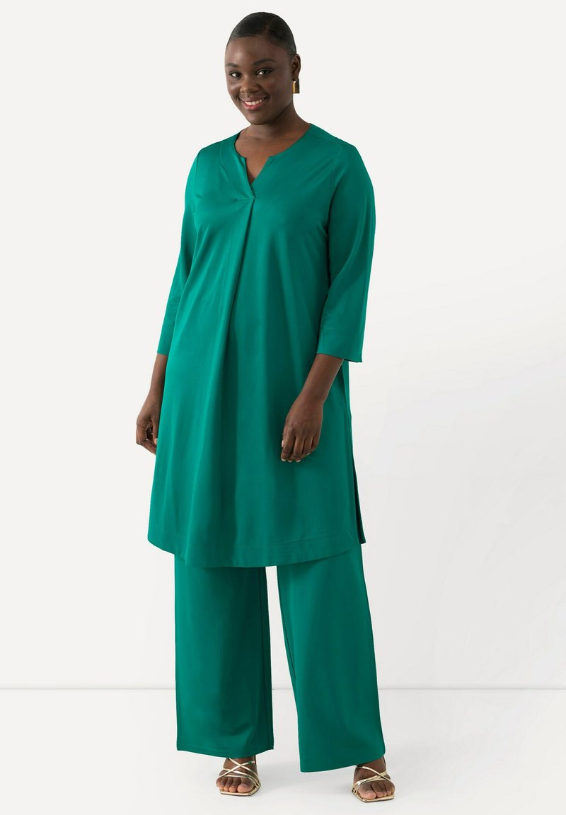 Teal, lång tunika med V-ringning och sidoöppningar, kombinerad med vida byxor. Slät, lättviktsmaterial med en avslappnad passform. Gula sandaletter med remmar.