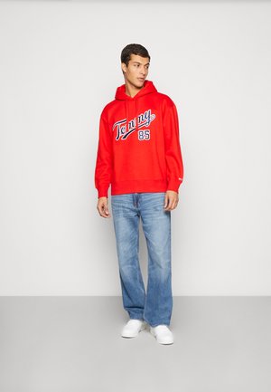 Sudadera de algodón roja con el logo "Tommy 85" en blanco y marino. Combinada con jeans de mezclilla azul claro y zapatillas blancas. Vista frontal plana.