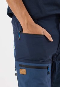 Marinblå utomhusbyxor med strukturerad yta, flera dragkedjefickor inklusive en framträdande sidoficka, samt en lädermärke med varumärket.