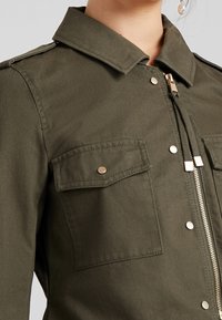 Oliwkowa kurtka z zapięciem na zamek, dwoma kieszeniami na piersiach z zatrzaskami oraz klasycznym kołnierzem z epauletami na ramionach.