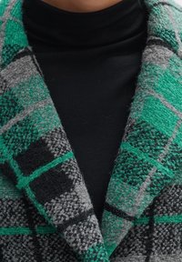 Nahaufnahme des Kragens eines grün-schwarz-grauen karierten Wollmantels über einem schwarzen hochgeschlossenen Oberteil.